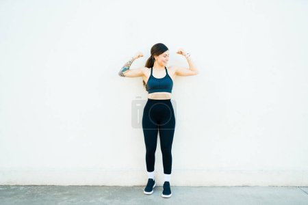 Téléchargez les photos : Forte femme en forme heureuse en vêtements de sport souriant faire des boucles biceps et l'exercice à l'extérieur avec beaucoup de force - en image libre de droit