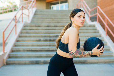 Foto de Hermosa mujer joven en ropa deportiva haciendo ejercicio de entrenamiento cruzado al aire libre y usando una bola de slam - Imagen libre de derechos