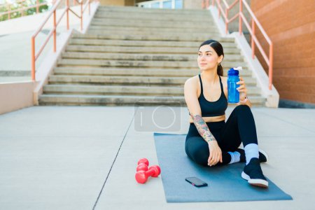 Photo for Smiling fit young woman with a water bottle looking happy about working out outdoors on an exercise mat - Royalty Free Image