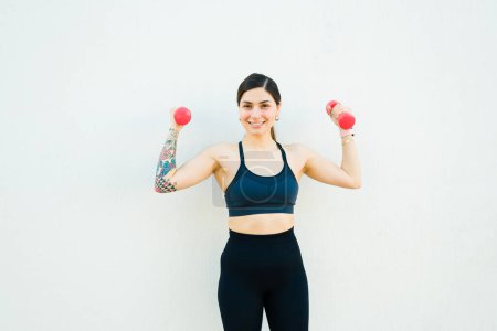 Photo for Athletic sporty fit woman smiling looking at the camera while lifting dumbbell weights during her strong workout - Royalty Free Image