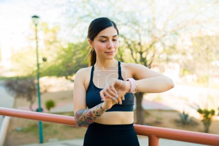Téléchargez les photos : Attrayant sportif jeune femme souriant tout en utilisant son application smartwatch et fitness lors de ses exercices d'entraînement à l'extérieur - en image libre de droit
