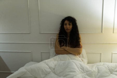 Photo for Angry latin woman with curly hair sitting in bed waking up in the middle of the night while suffering from insomnia - Royalty Free Image