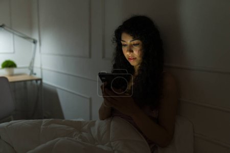 Photo for Beautiful latin woman texting on her smartphone late at night before sleeping or suffering from insomnia in the bedroom - Royalty Free Image