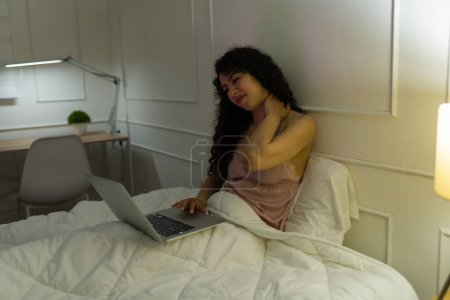 Foto de Mujer joven cansada mirando estresada y sufriendo de dolor de cuello antes de dormir mientras usa su computadora portátil en la cama - Imagen libre de derechos