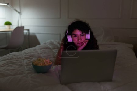 Téléchargez les photos : Jeune femme détendue avec écouteurs regardant un film sur l'ordinateur portable tout en se relaxant au lit en mangeant du pop-corn - en image libre de droit