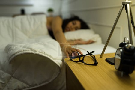 Téléchargez les photos : Latino jeune femme se réveillant dans son lit et atteignant sa table de chevet pour mettre ses lunettes le matin - en image libre de droit
