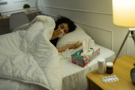 Photo for Sick latin young woman under the covers resting in bed and blowing her nose while suffering from a bad cold - Royalty Free Image