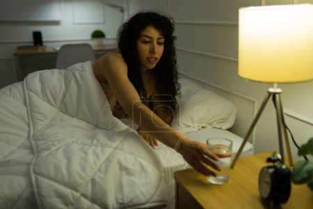 Photo for Thirsty hispanic woman with curly hair waking up in bed and drinking a glass of water in the middle of the night - Royalty Free Image