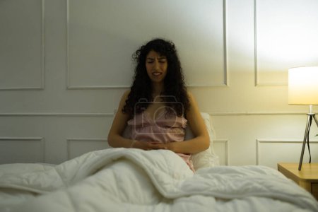 Téléchargez les photos : Malade inquiet jeune femme souffrant de maux d'estomac ou des douleurs de crampes tout en se reposant dans le lit sous les couvertures - en image libre de droit