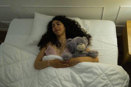 Téléchargez les photos : Vue de dessus d'une femme latine heureuse souriante regardant détendue et étreignant un ours en peluche tout en souriant et en dormant - en image libre de droit