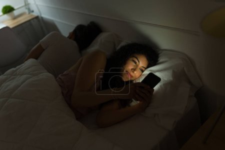 Téléchargez les photos : Joyeux jeune femme couchée debout la nuit et souriant tout en envoyant des SMS sur le smartphone tout en couchant avec son partenaire - en image libre de droit