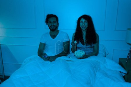 Foto de Atractiva pareja joven feliz en pijama comiendo palomitas de maíz en la cama mientras ven una divertida película de comedia juntos por la noche - Imagen libre de derechos