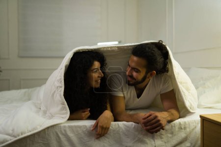 Téléchargez les photos : Couple affectueux joyeux amoureux souriant tout en se blottissant sous les couvertures au lit tout en se préparant à dormir - en image libre de droit