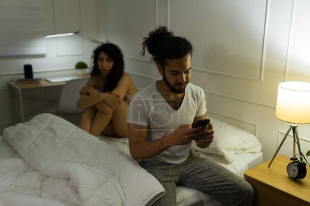 Téléchargez les photos : Mensonge hispanique homme textos sur le smartphone tout en trichant sur sa triste petite amie se préparant à dormir dans la chambre - en image libre de droit