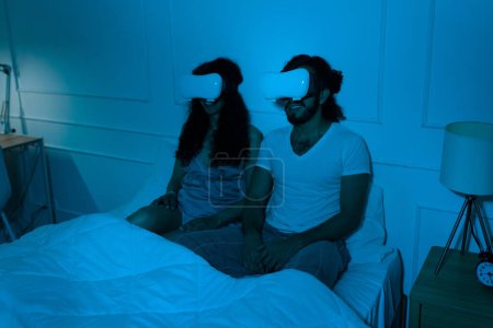 Téléchargez les photos : Jeune couple amusant portant des lunettes de réalité virtuelle et regardant une vidéo immersive assis au lit la nuit - en image libre de droit