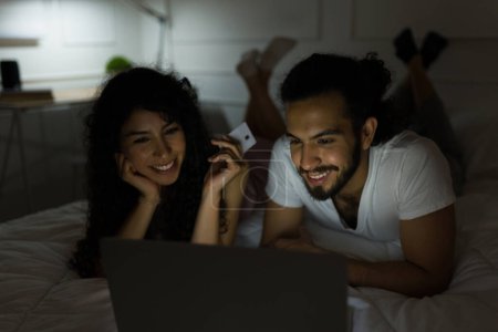 Foto de Atractiva pareja joven sonriendo en la cama mientras disfruta de las compras en línea en el portátil y comprar con una tarjeta de crédito - Imagen libre de derechos