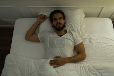 Téléchargez les photos : Agacé hispanique jeune homme couché éveillé la nuit tout en étant couché en essayant de dormir tout en souffrant d'insomnie - en image libre de droit