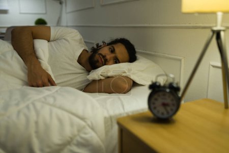 Photo for Distressed hispanic man lying awake late at night and watching the alarm clock while suffering from insomnia - Royalty Free Image
