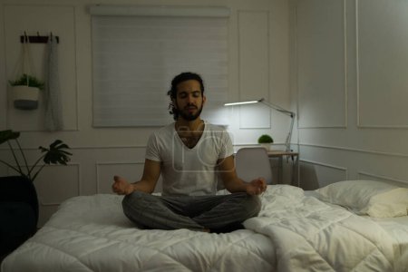 Foto de Hombre hispano relajado en pijama escuchando música relajante con auriculares mientras hace una meditación en la cama antes de dormir - Imagen libre de derechos