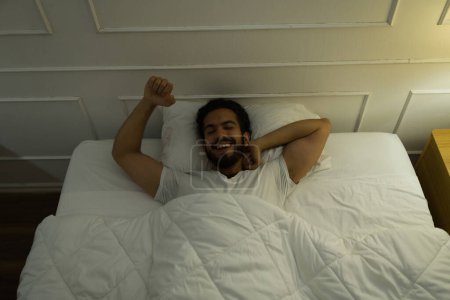Photo for Happy attractive young man smiling and feeling relaxed and well rested after waking up and stretching in bed - Royalty Free Image