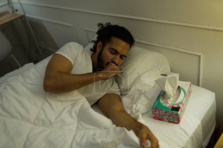 Téléchargez les photos : Jeune homme hispanique tousse et souffre d'un rhume malade en utilisant des tissus et couché éveillé dans son lit - en image libre de droit