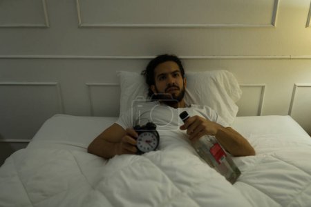Foto de Guapo joven que tiene problemas para dormir debido a su adicción al alcohol y beber acostado en la cama - Imagen libre de derechos