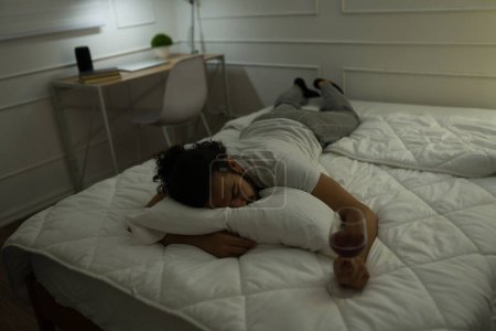Téléchargez les photos : Jeune homme épuisé avec une dépendance à l'alcool dormant seul au lit avec un verre de vin après avoir bu - en image libre de droit