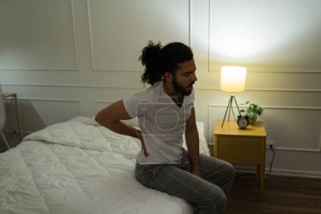 Foto de Hombre hispano estresado en ropa de dormir despertando de la cama con dolor lumbar debido al mal sueño - Imagen libre de derechos