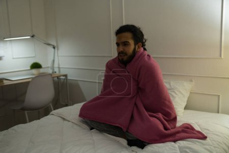 Téléchargez les photos : Jeune homme bouleversé enveloppé dans une couverture et couvrant du froid au lit pendant l'hiver ayant des problèmes de sommeil - en image libre de droit