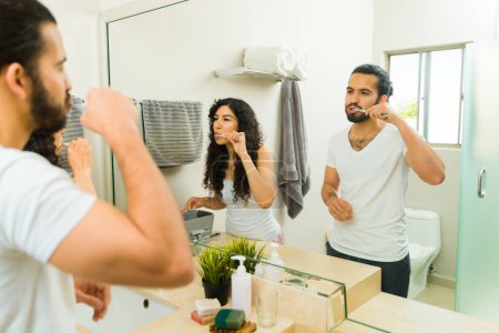 Foto de Pareja joven latina mirándose juntos en el espejo del baño mientras se cepillan los dientes durante su rutina matutina - Imagen libre de derechos