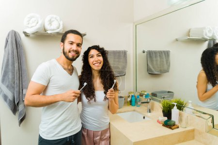 Foto de Pareja mexicana atractiva sonriendo en pijama durante la mañana mientras se cepillan los dientes en el baño - Imagen libre de derechos