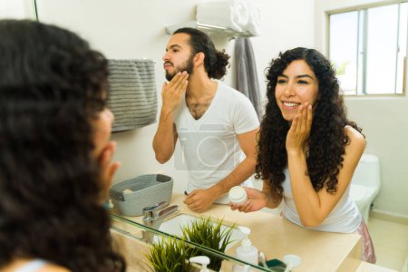 Téléchargez les photos : Couple heureux hispanique souriant tout en utilisant des produits de soins de la peau et se préparer le matin ensemble dans la salle de bain - en image libre de droit