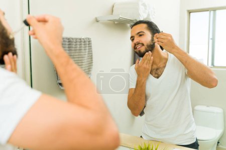 Foto de Alegre hombre atractivo usando aceite hidratante en su barba y aseo en el espejo del baño - Imagen libre de derechos