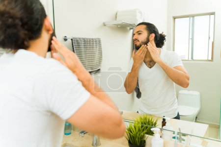 Foto de Hombre hispano guapo en el baño arreglando su barba y usando productos de limpieza en la mañana - Imagen libre de derechos