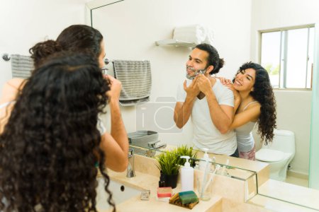 Photo for Cheerful young man shaving his beard and talking with his girlfriend while getting ready in the morning - Royalty Free Image