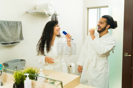 Foto de Alegre pareja latina divirtiéndose durante su rutina de limpieza matutina y cantando en el baño escuchando música - Imagen libre de derechos