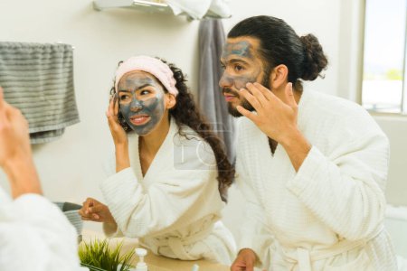 Téléchargez les photos : Femme hispanique et homme portant un masque visage au charbon actif pour exfolier leur peau lors d'un spa à domicile - en image libre de droit