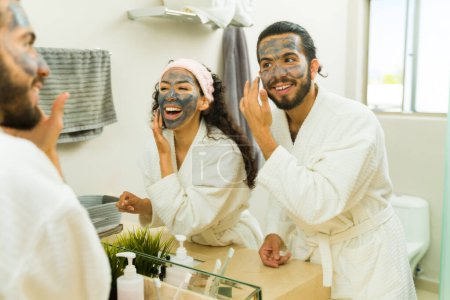 Foto de Emocionada joven pareja riendo divirtiéndose mientras se pone la máscara de barro facial y haciendo su rutina de cuidado de la piel juntos - Imagen libre de derechos