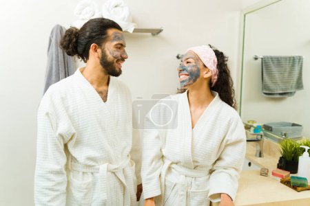 Foto de Emocionada hermosa pareja exfoliando su piel usando máscaras de carbón activado juntos usando túnicas blancas cómodas - Imagen libre de derechos