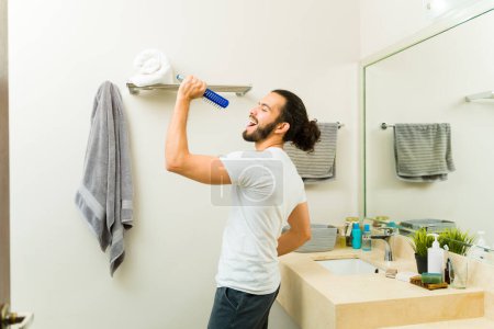Foto de Emocionado hombre feliz divirtiéndose mientras se prepara en el baño y cantando con un cepillo de pelo durante la mañana - Imagen libre de derechos