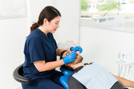 Téléchargez les photos : Dentiste féminine en blouse bleue souriant en faisant un examen dentaire sur un jeune homme et en vérifiant les cavités - en image libre de droit