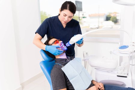 Foto de Profesional mujer caucásica dentista haciendo un tratamiento con láser blanqueador en un hombre joven para los dientes blancos - Imagen libre de derechos