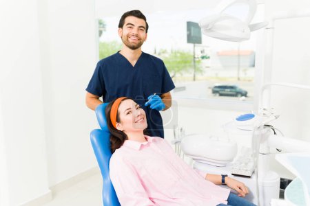 Photo for Hispanic male doctor smiling while treating a young woman at the beauty clinic getting a filling procedure or hyaluronic acid injections - Royalty Free Image