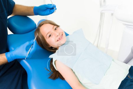 Foto de Adorable joven sonriendo haciendo contacto visual mientras se ve feliz durante un examen dental en el consultorio del dentista - Imagen libre de derechos