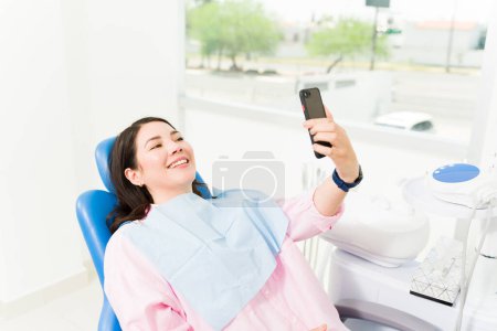 Téléchargez les photos : Femme caucasienne souriant regardant heureux de prendre un selfie avec un smartphone pour poster sur les médias sociaux tandis que chez le dentiste pour un examen - en image libre de droit