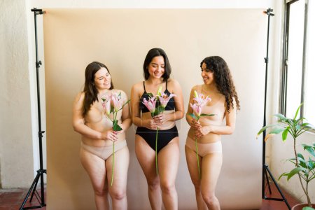 Téléchargez les photos : Attrayant diverses femmes féminines en sous-vêtements tenant des fleurs dans le studio avec beaucoup de positivité du corps - en image libre de droit