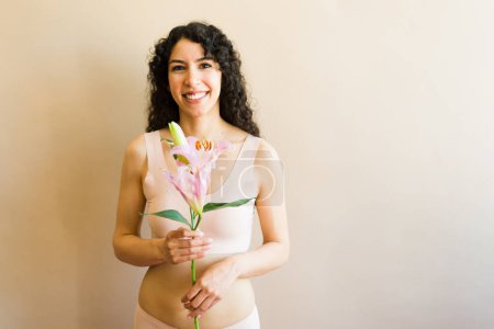 Téléchargez les photos : Latin femme heureuse avec les cheveux bouclés souriant sur l'acceptation du corps et l'amour de soi dans ses sous-vêtements tout en tenant des fleurs féminines - en image libre de droit