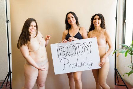 Foto de Felices hermosas amigas en ropa interior sosteniendo un cartel de positividad corporal y abrazando el amor propio femenino - Imagen libre de derechos