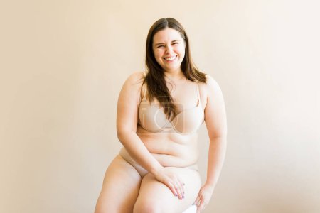 Téléchargez les photos : Portrait d'une jolie grosse femme joufflue souriante qui semble heureuse en étant assise dans ses sous-vêtements en contact visuel - en image libre de droit