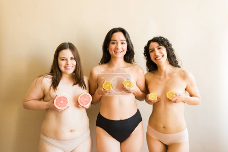 Téléchargez les photos : Trois jeunes femmes diversifiées avec leur poitrine nue mettant des fruits orange sur leur poitrine et se sentant confiantes au sujet de leurs corps diversifiés - en image libre de droit